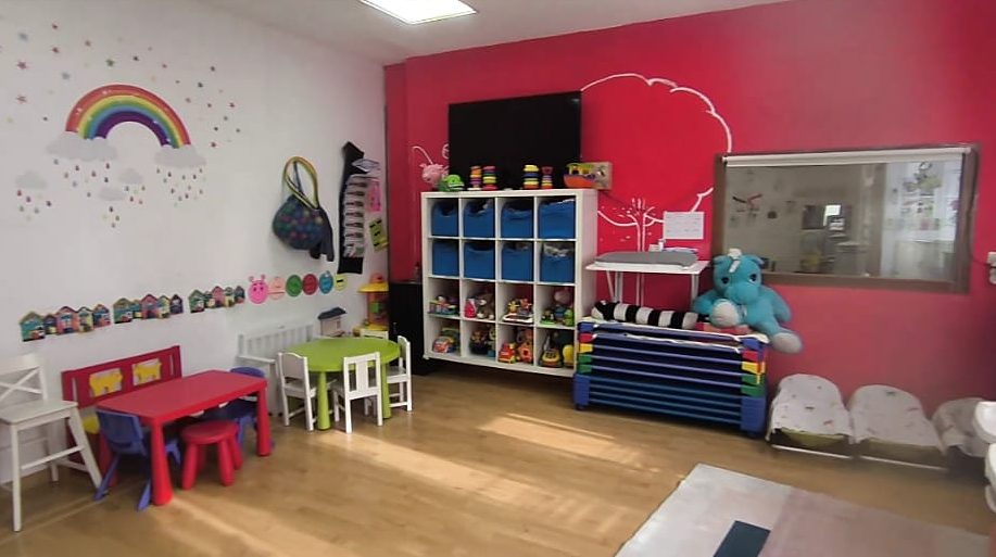 Aula Arcoíris con pared roja, estantería de juguetes, mesitas de colores y hamacas para la siesta