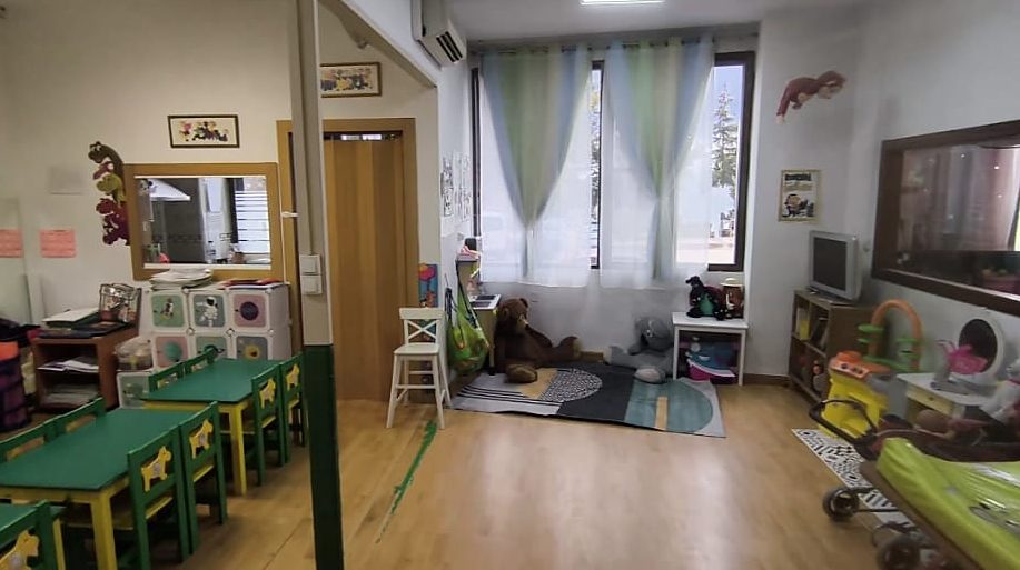 Aula Verde con mesitas verdes y amarillas, rincón de lectura con peluches y grandes ventanales