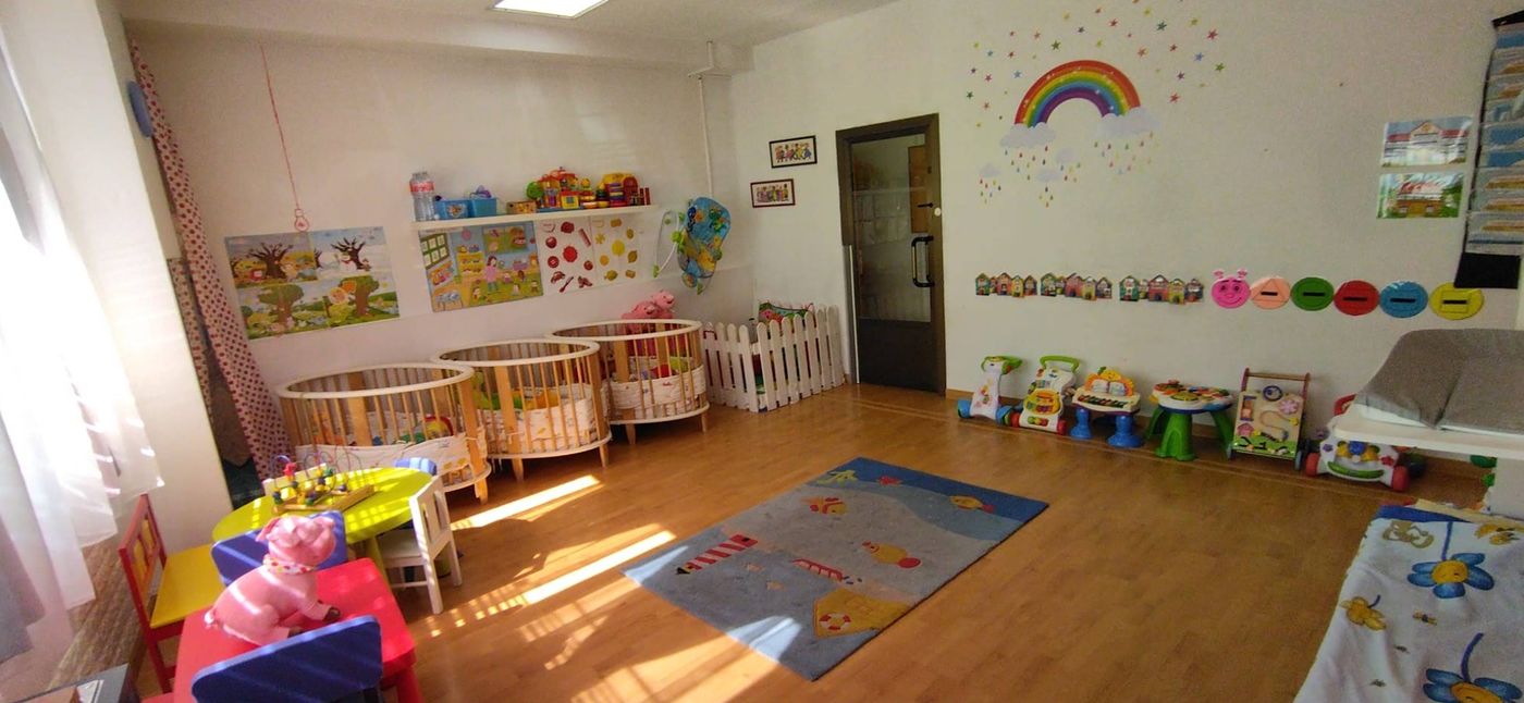 Aula de bebés con tres cunas de madera, valla blanca, andadores, alfombra infantil y un arcoíris pintado en la pared