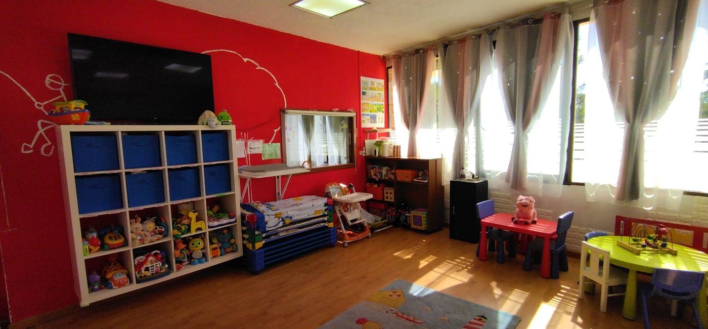 Aula con pared roja, estantería de juguetes, hamacas apiladas, mesitas de colores y grandes ventanales