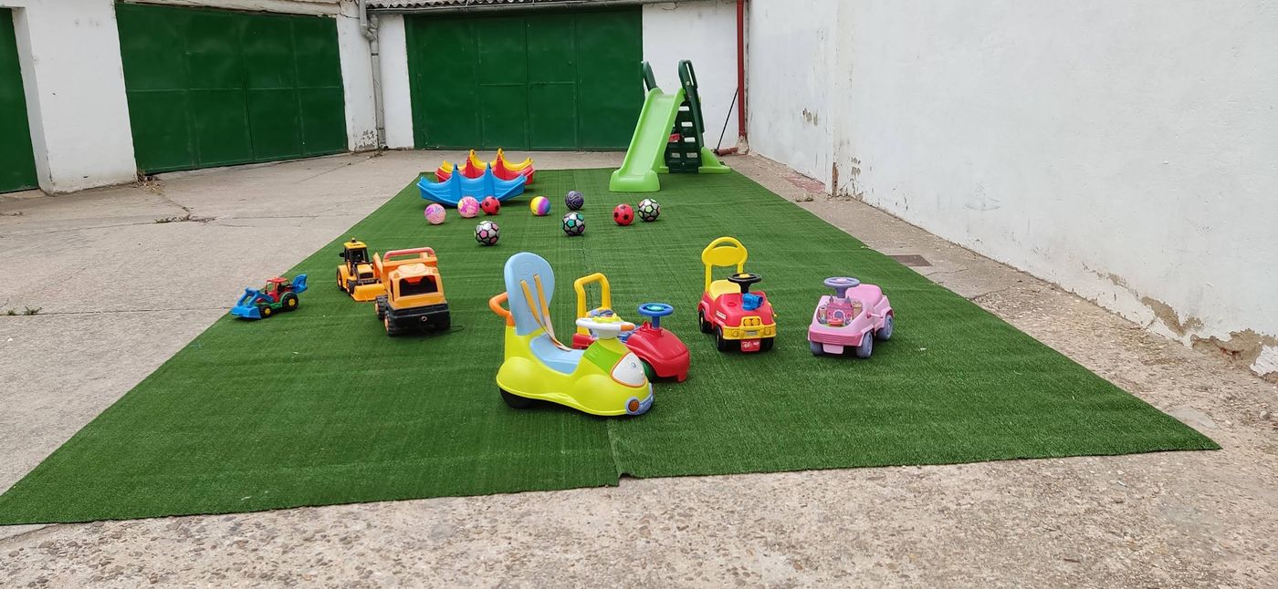 Patio exterior con césped artificial, tobogán verde, balancines, pelotas de colores y correpasillos